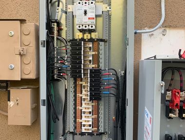 Open electrical panel with circuit breakers and wiring beside outdoor utility boxes.