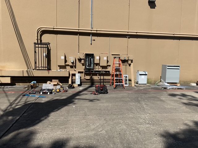 Outdoor utility wall with pipes, meters, and equipment, plus a ladder and scattered tools on asphalt.