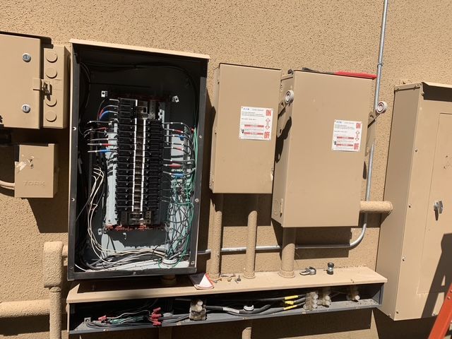 Open electrical panel with exposed wiring and gray utility boxes on a beige wall.