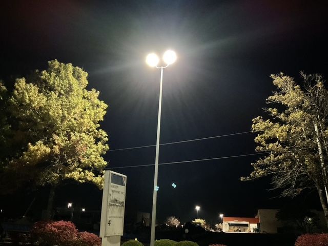 Nighttime parking lot with bright streetlights, trees, and a sign against a dark sky.