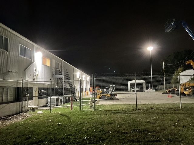 Nighttime view of a fenced industrial yard with lit building, grass, and equipment under bright lights