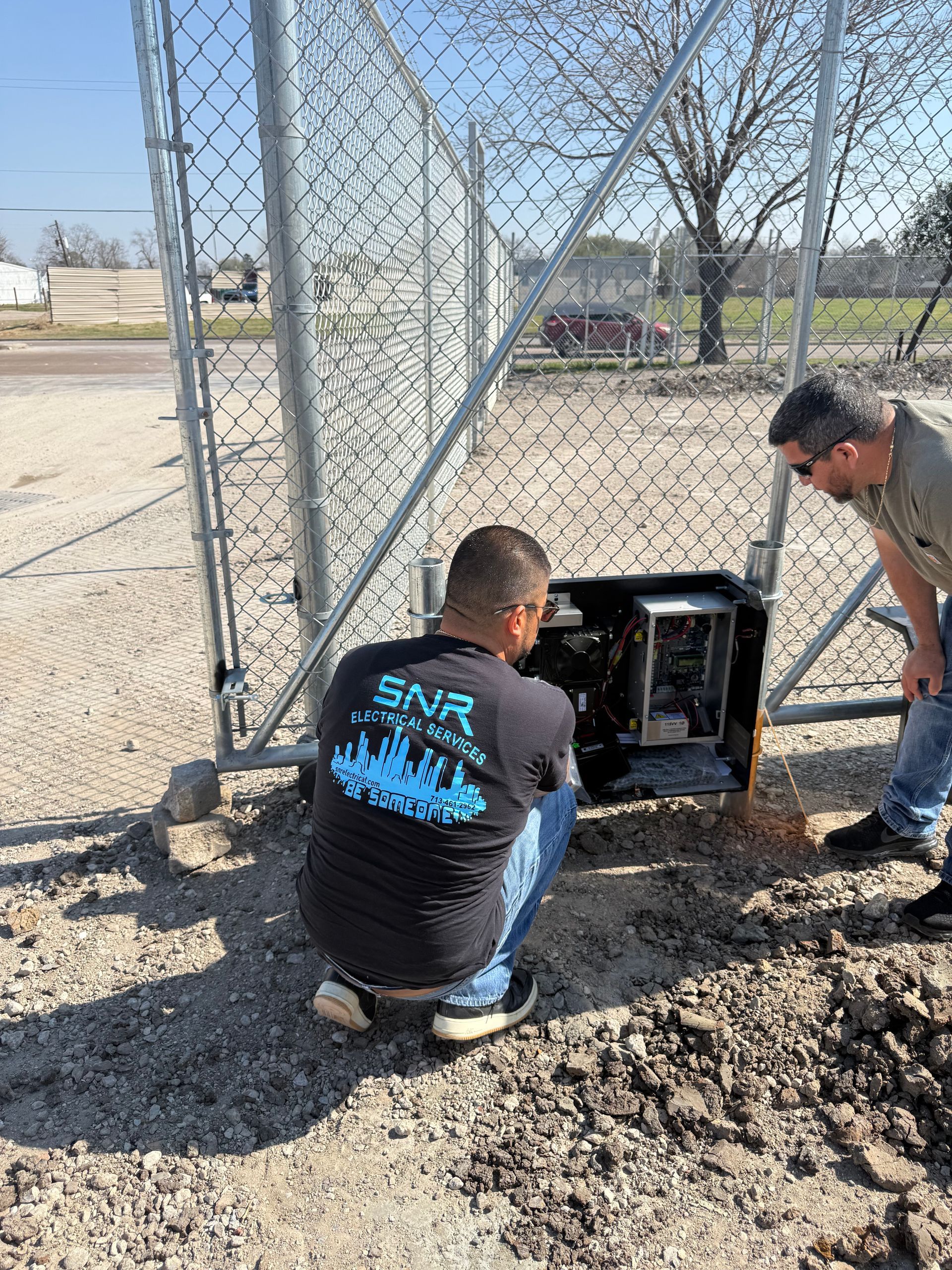 Two workers inspect an open utility box beside a chain-link fence in a gravel yard.
