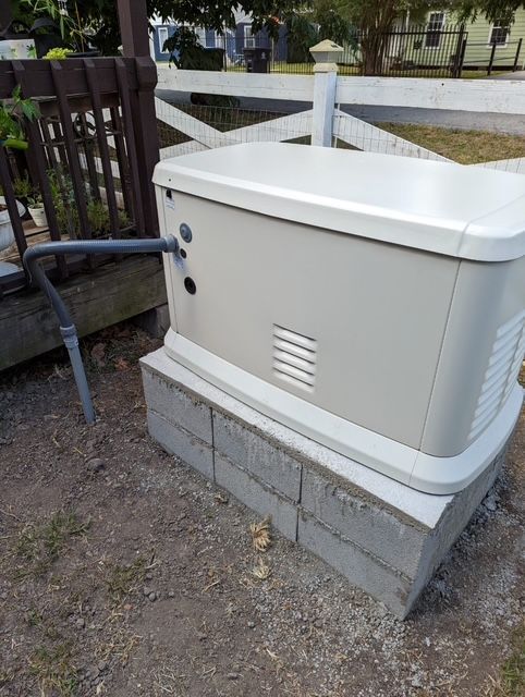White outdoor generator beside a fence, mounted on a cinder block base near a house yard