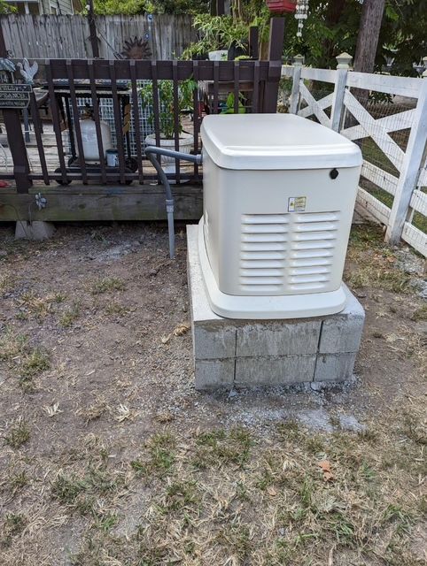 White outdoor generator on cinder blocks beside a fence and deck in a gravel yard.
