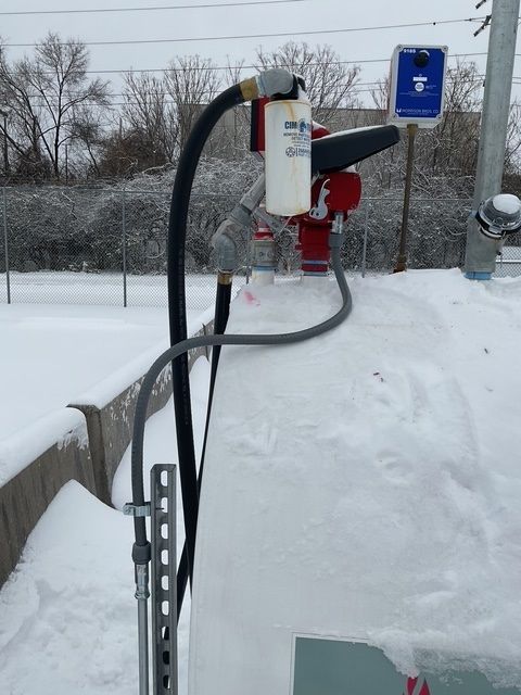 Snow-covered roadside emergency pump station with hoses and a blue safety sign.