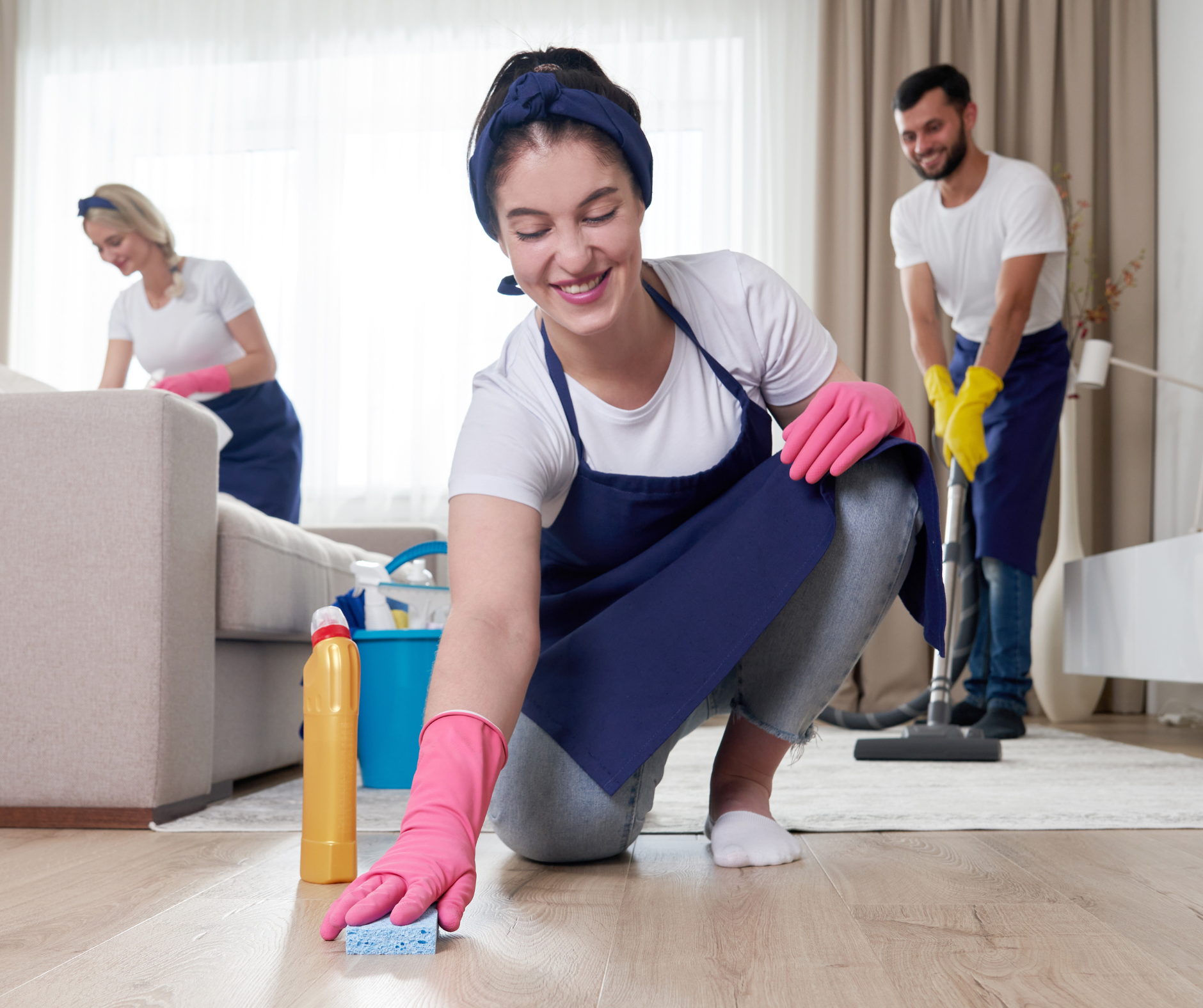 Three people cleaning a room. A woman squats scrubbing a floor, smiling. A man vacuums, another woman dusts.