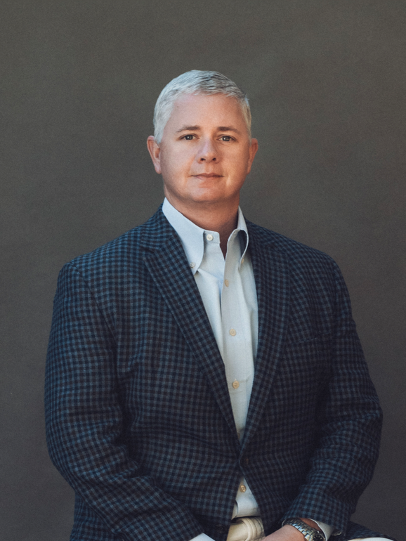 Jack Daniels in a suit and blue shirt is standing in front of a wooden wall.