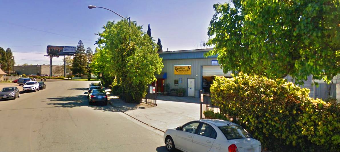 Exterior of a car repair shop with a billboard in the background, parked cars, and trees.