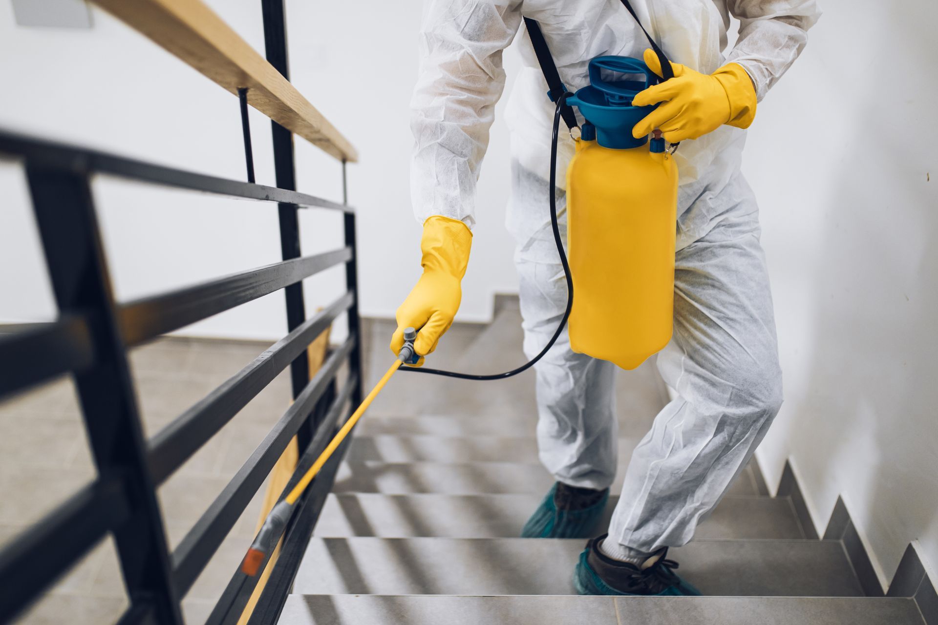 A person in protective gear disinfects a staircase handrail with a yellow handheld sprayer.