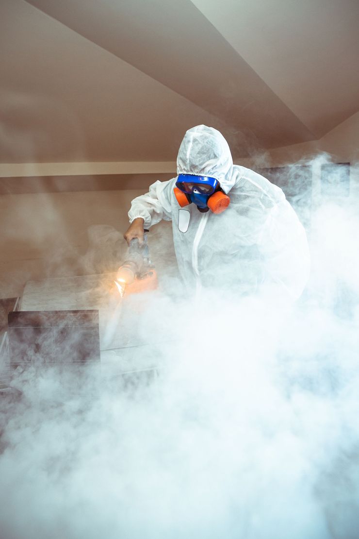 A person in a white hazmat suit and respirator mask stands amidst thick white smoke in an indoor room.