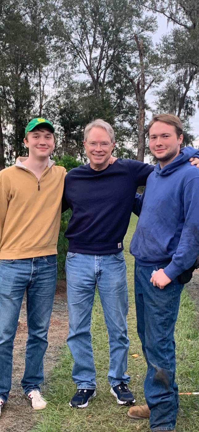 David E. Hill and his sons standing together outdoors on grass, smiling with trees in the background.