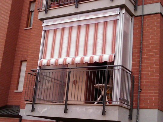 Balcone con edificio in mattoni rossi, tenda a strisce, ringhiera nera e tavolo all'interno.