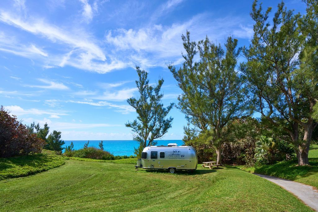 Silver camper trailer on grassy seaside lawn with blue ocean and trees under a bright sky