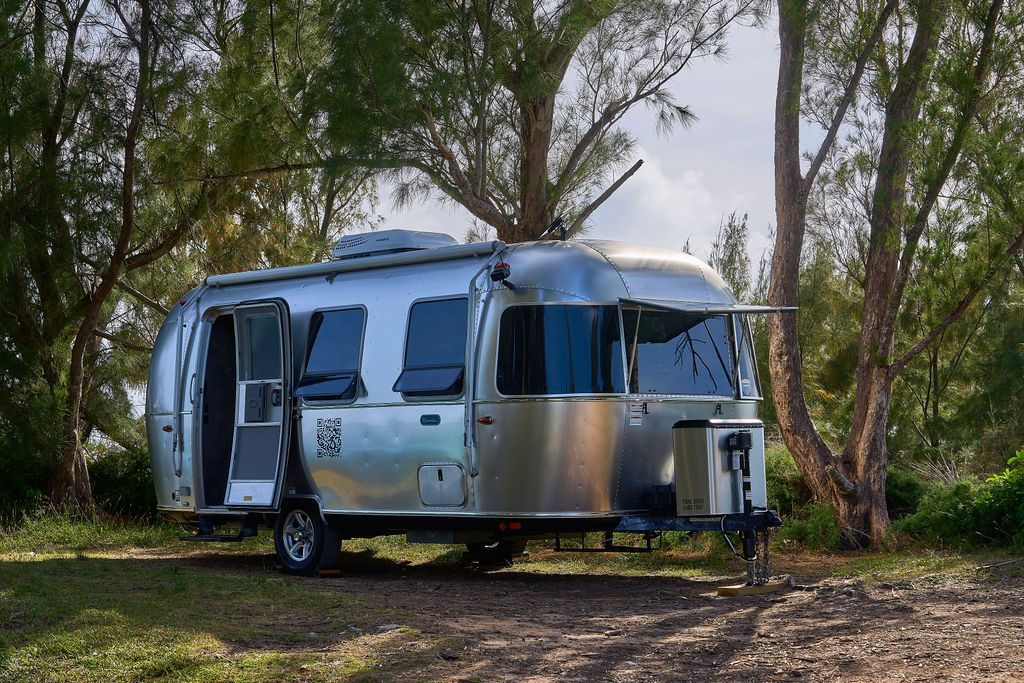 Silver camper trailer parked among trees in a wooded area with its door open, Coney Island