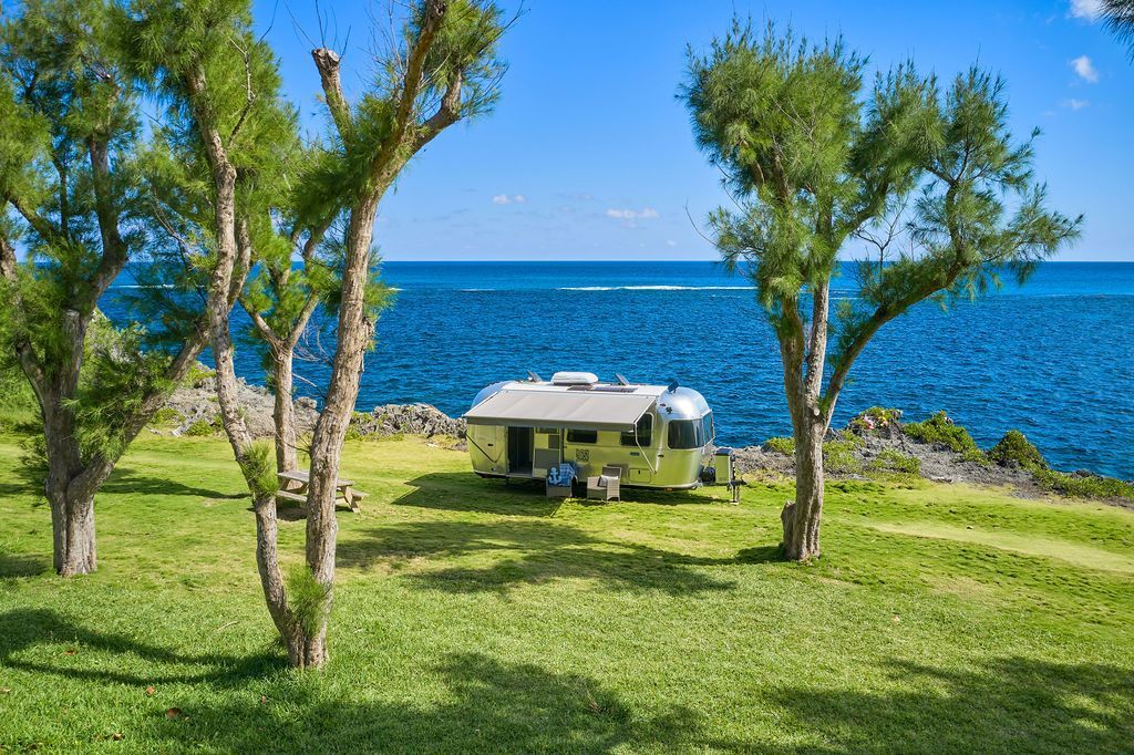 Green grassy seaside yard with trees and a Silver RV overlooking a blue ocean