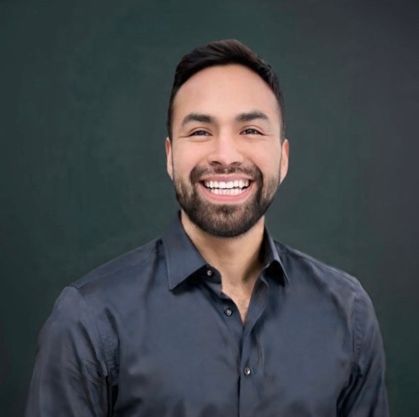 A smiling person with a beard, wearing a dark gray button-down shirt against a dark green background.