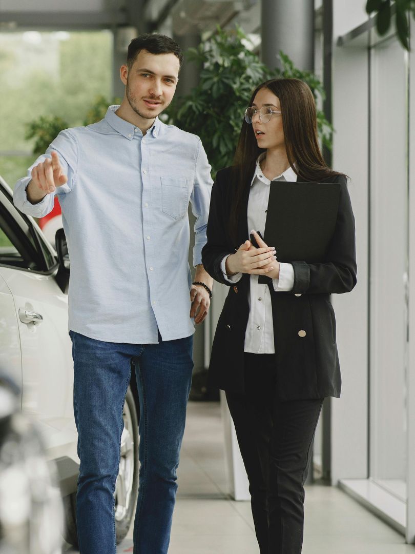 Two professionals walking indoors beside a car, one pointing ahead while the other holds a folder.