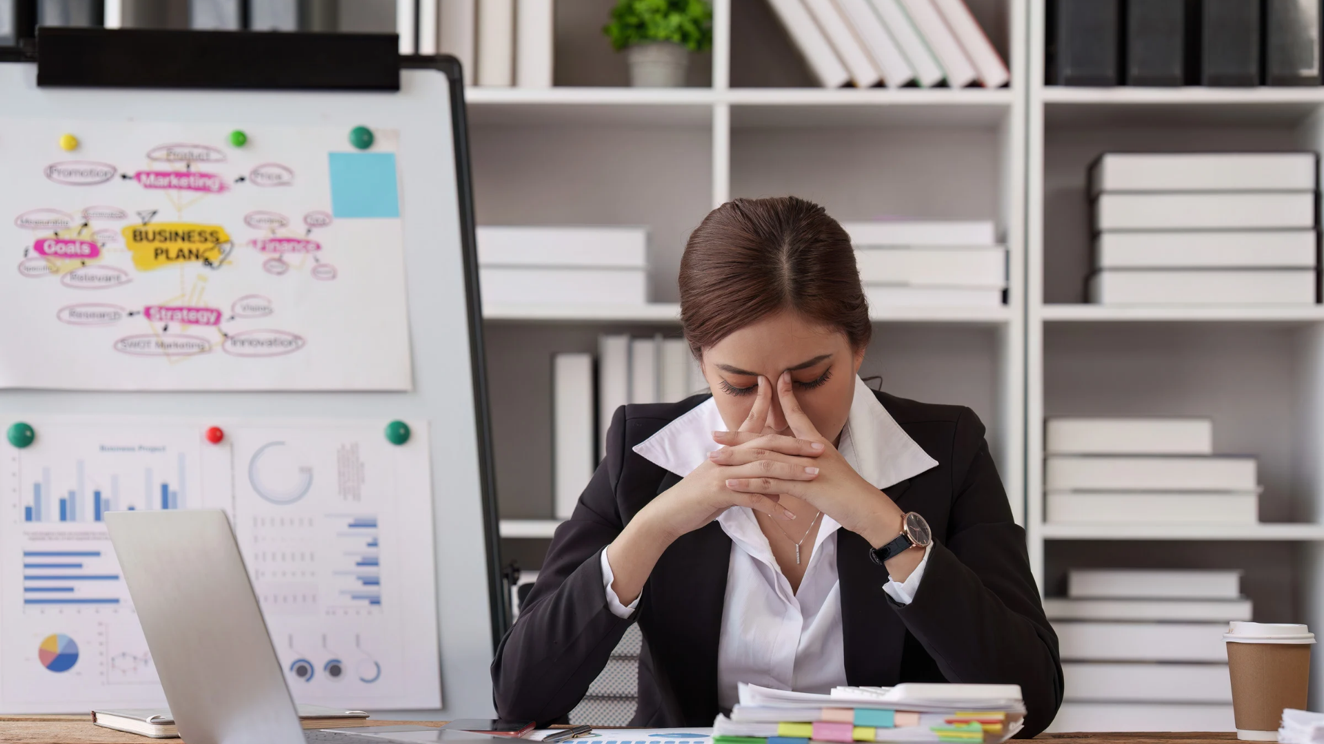 Person sitting at a desk, head in hands, with charts and notes on a whiteboard behind them