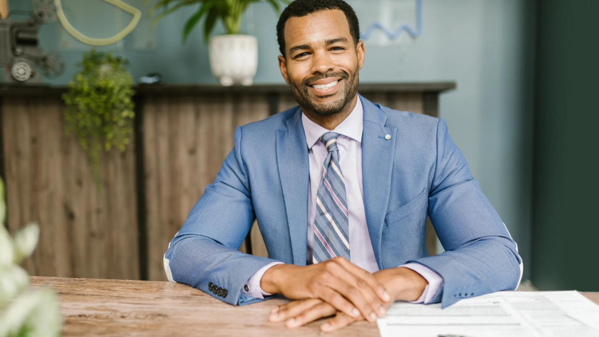 Smiling man in a blue blazer and tie seated at a desk with papers in a modern office setting