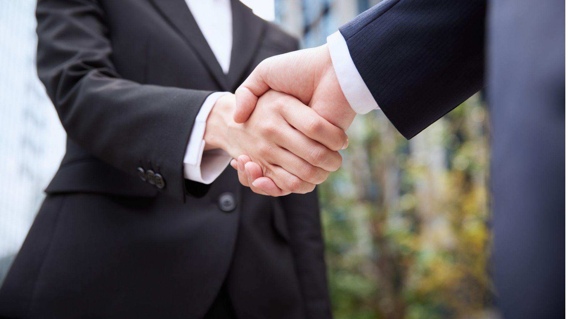 Two people in dark suits shaking hands outdoors, symbolizing a business agreement.