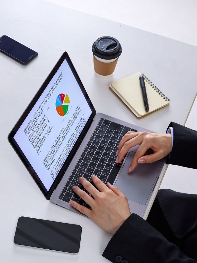 Hands typing on a laptop at a desk with notebook, coffee cup, and phone nearby.