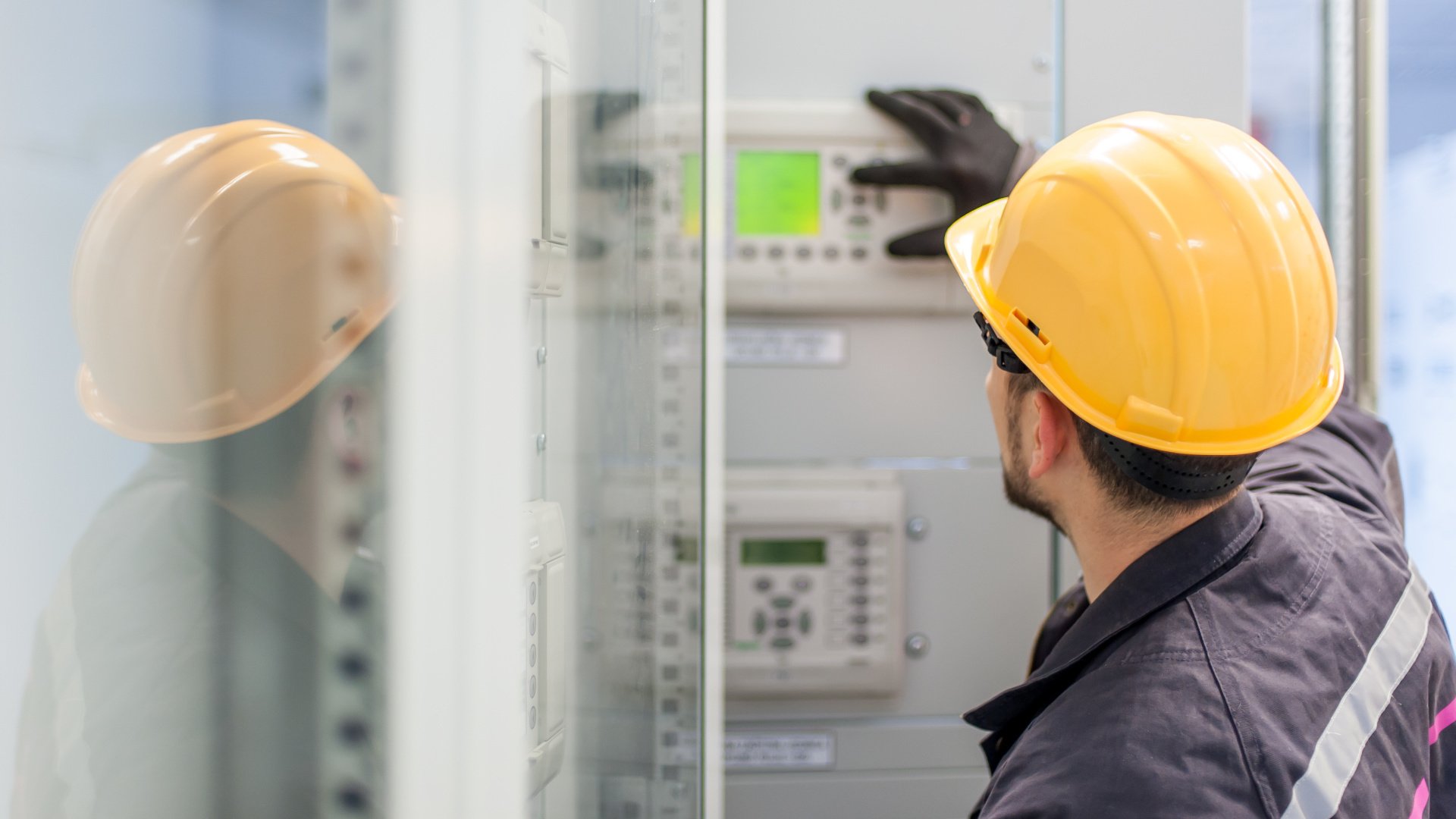 Un operaio con elmetto giallo e guanti neri ispeziona un pannello di controllo digitale all'interno di un armadio industriale.