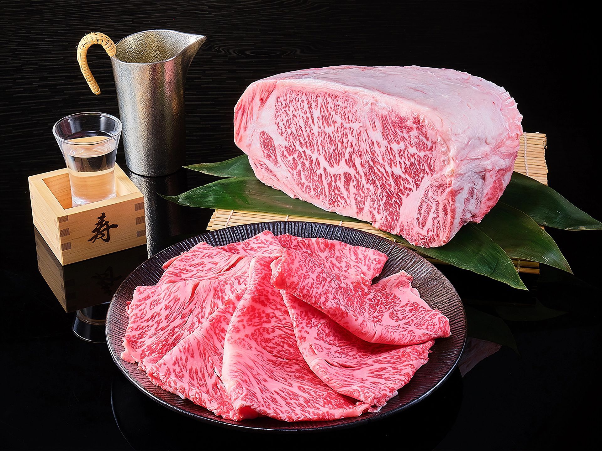 Slices of marbled Wagyu beef on a dark brown plate, with a large cut on green leaves and a bamboo mat in the background, accompanied by a sake cup and pitcher.