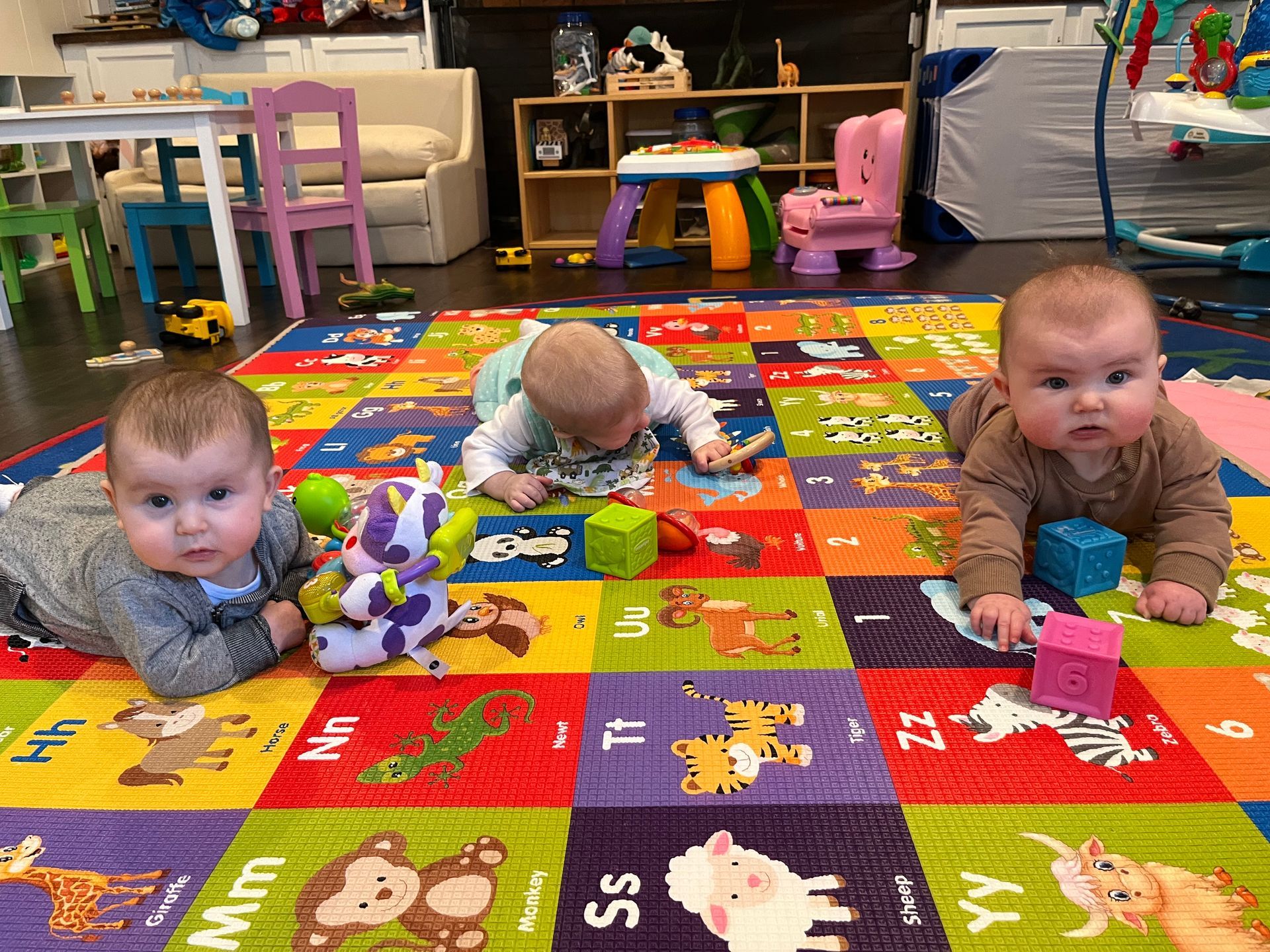 Three babies are laying on their stomachs on a colorful rug in a play room.