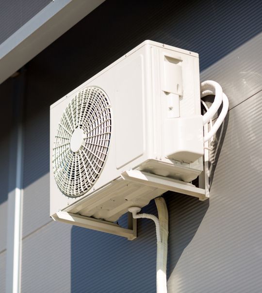 Unidade externa de ar condicionado montada na parede sob uma cobertura, com tubulações e fiação visíveis.