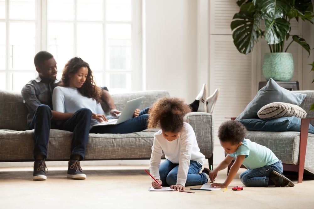Uma família está sentada em um sofá em uma sala de estar enquanto duas crianças brincam no chão.