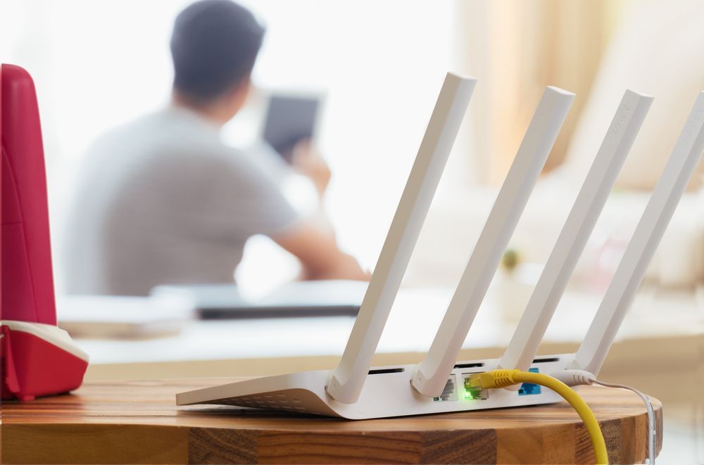 Um roteador está sobre uma mesa de madeira com um homem usando um tablet ao fundo.