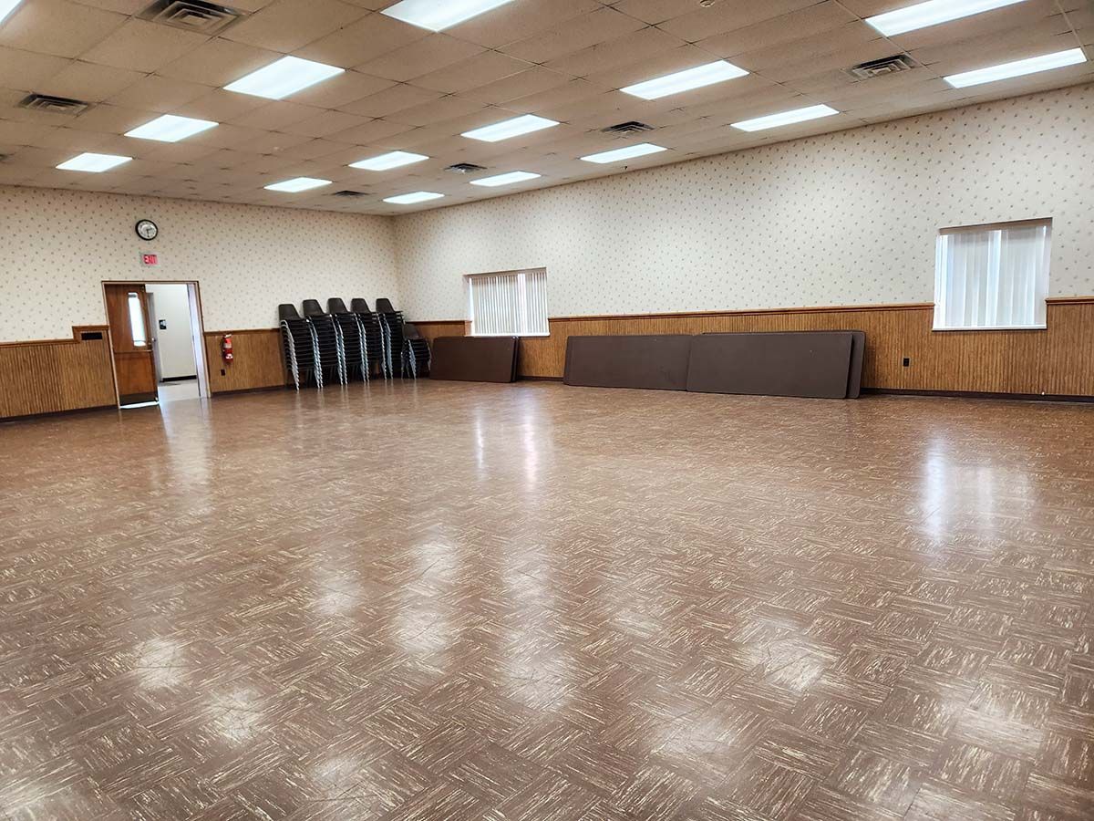 Empty community hall with polished floor, fluorescent lights, stacked chairs, and a wall table along one side