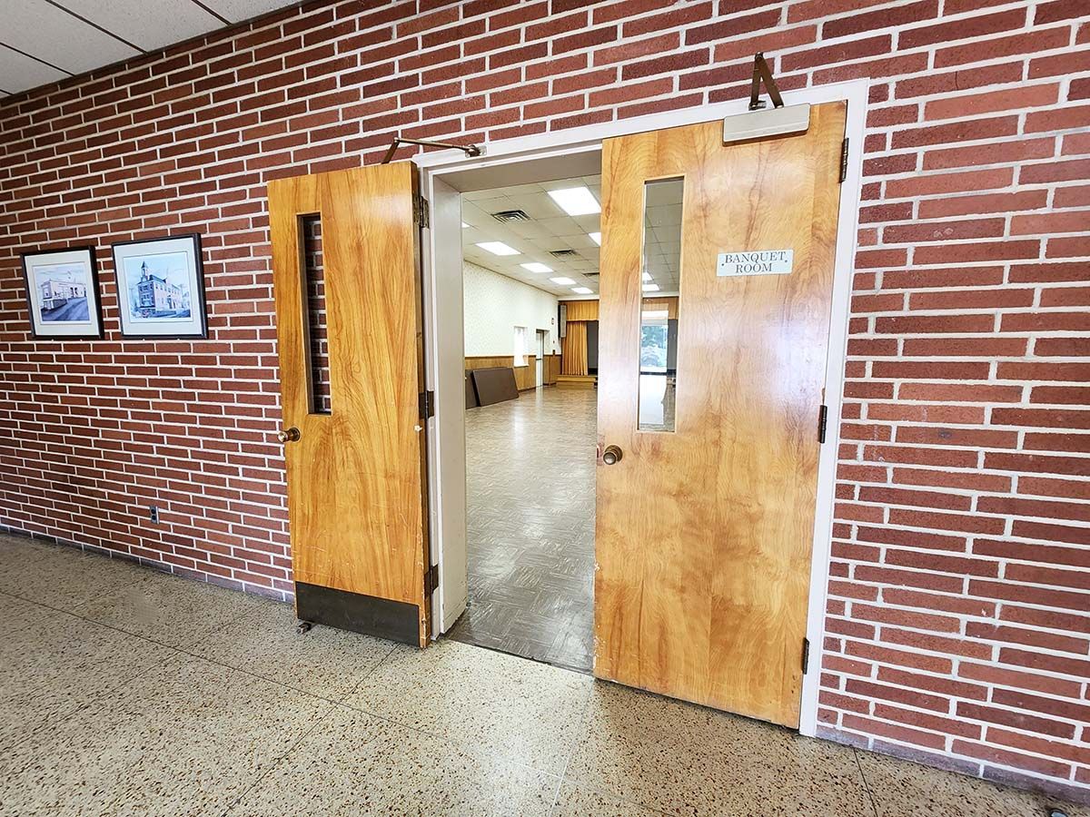 Open wooden double doors in a brick hallway, revealing a lit interior corridor.