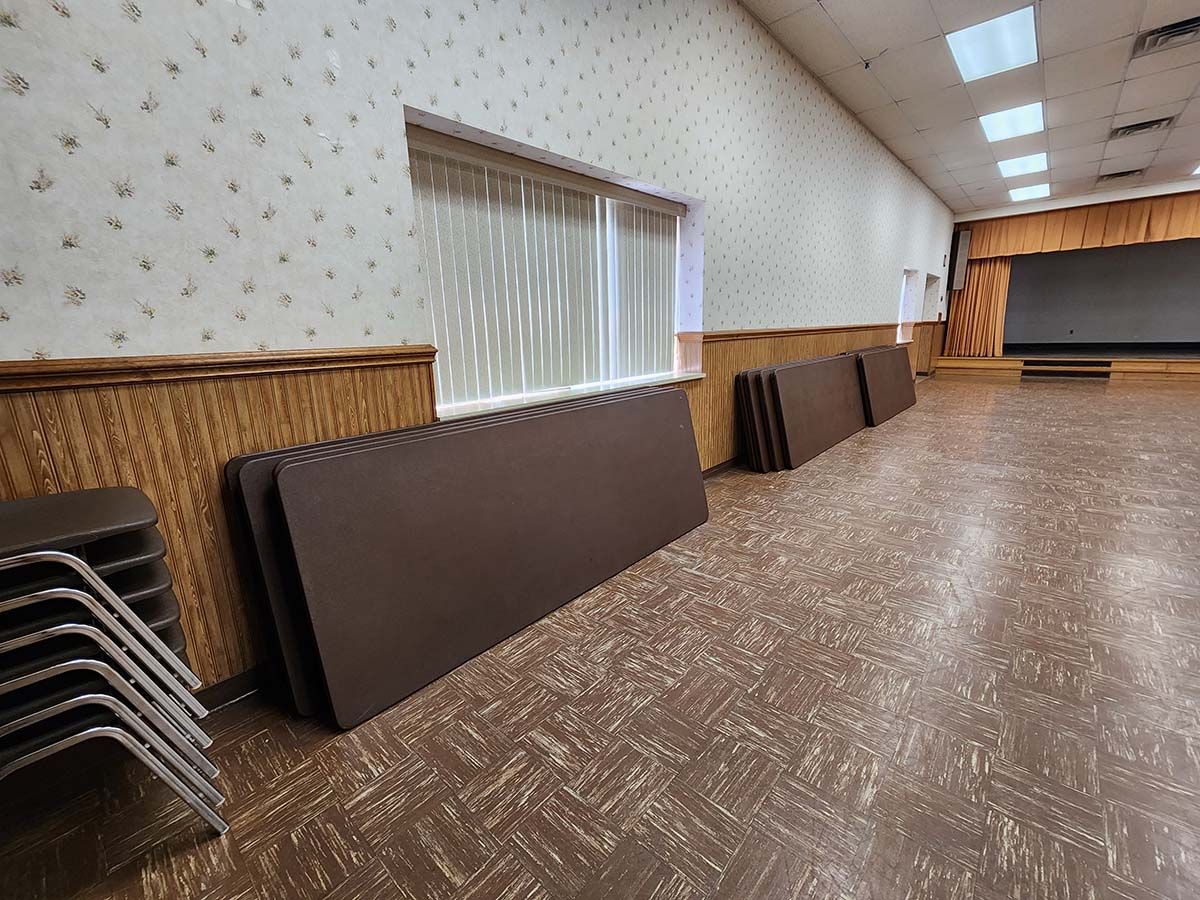 Empty banquet hall with stacked folding tables and chairs along the wall, brown patterned carpet, and stage at the end.