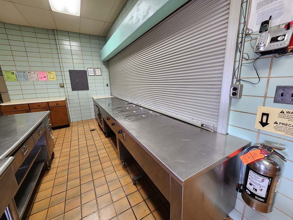Commercial kitchen with stainless steel counters, tiled walls, and a closed metal roll-up window.