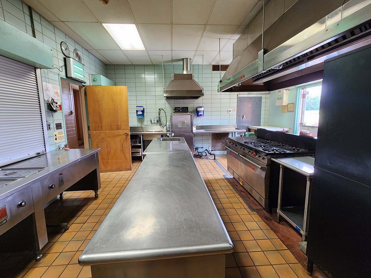 Empty commercial kitchen with stainless steel prep table, stovetop, cabinets, and hood vents