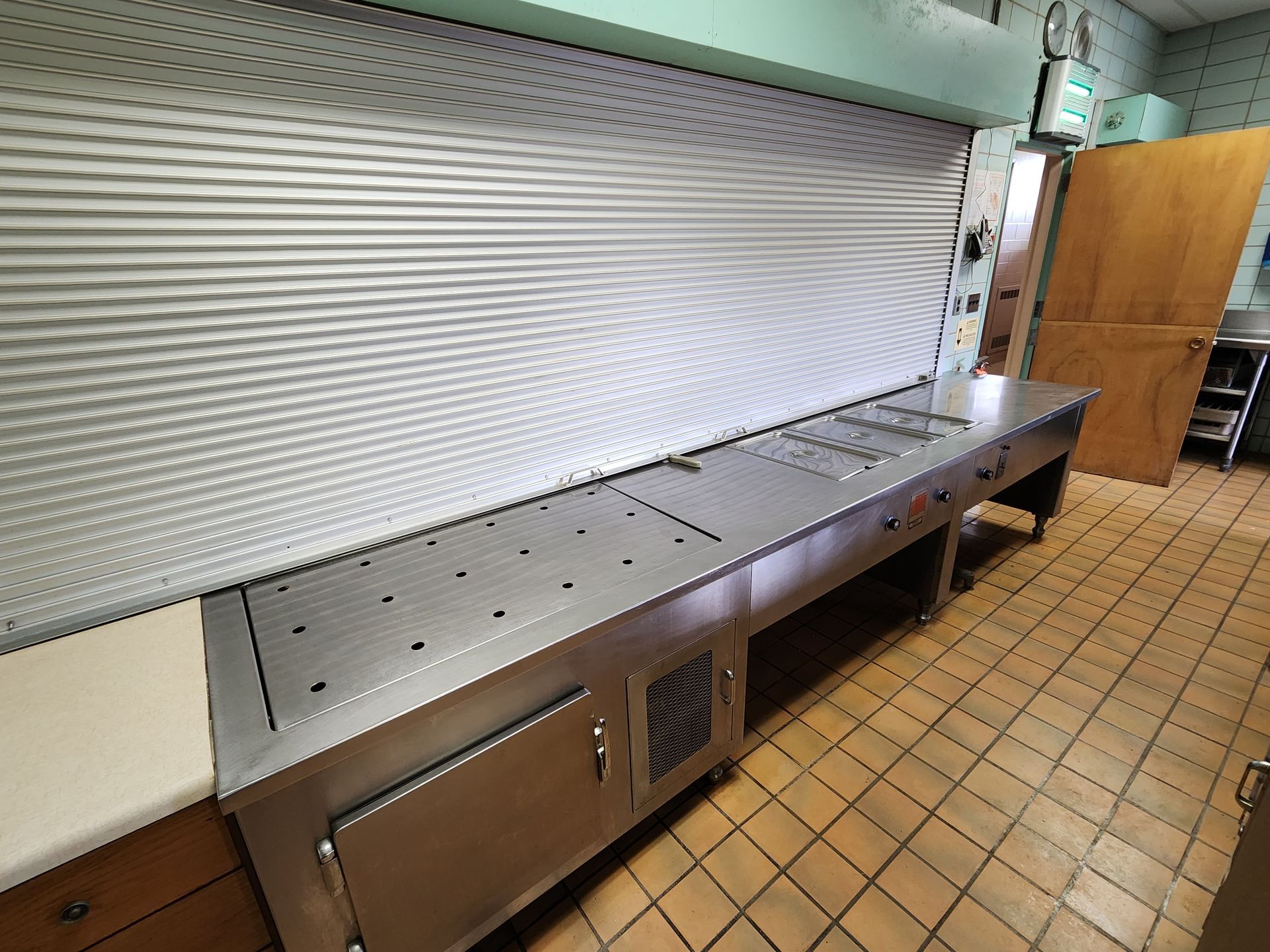 Stainless steel kitchen prep counter beneath a closed metal service window in a tiled room