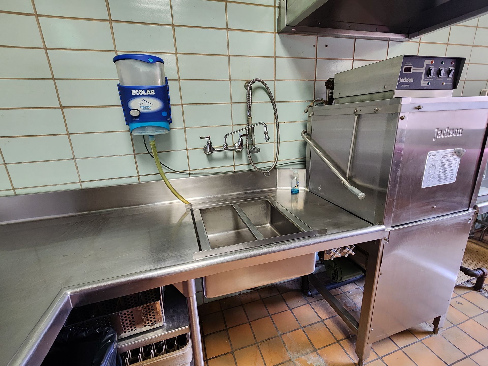 Commercial kitchen sink and stainless steel prep area with tiled wall and equipment.