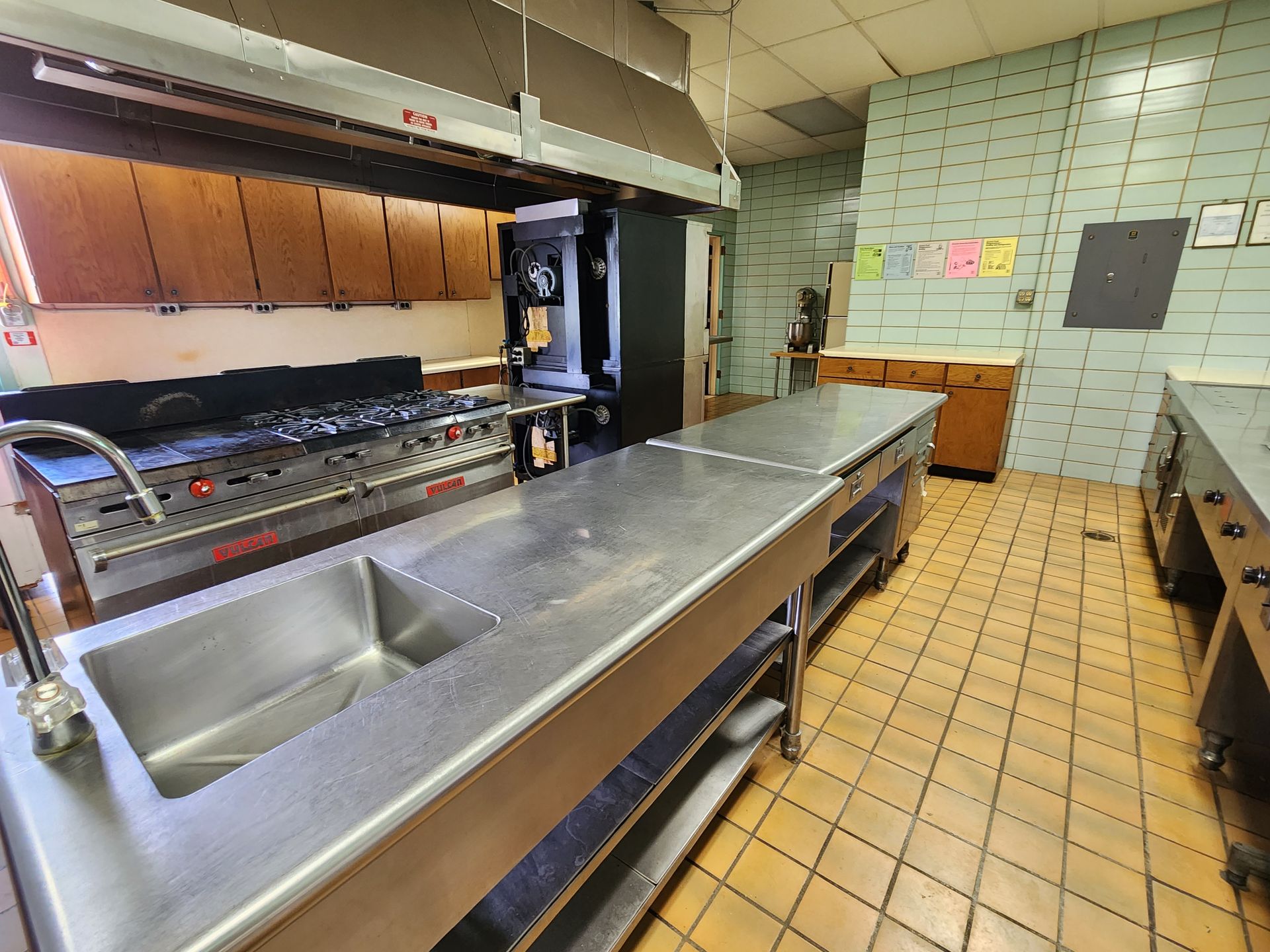 Empty commercial kitchen with stainless prep counter, sinks, and tiled floor.