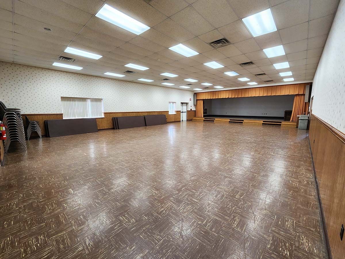 Empty indoor hall with fluorescent lights, wood floor, stage curtain, and tables along the walls