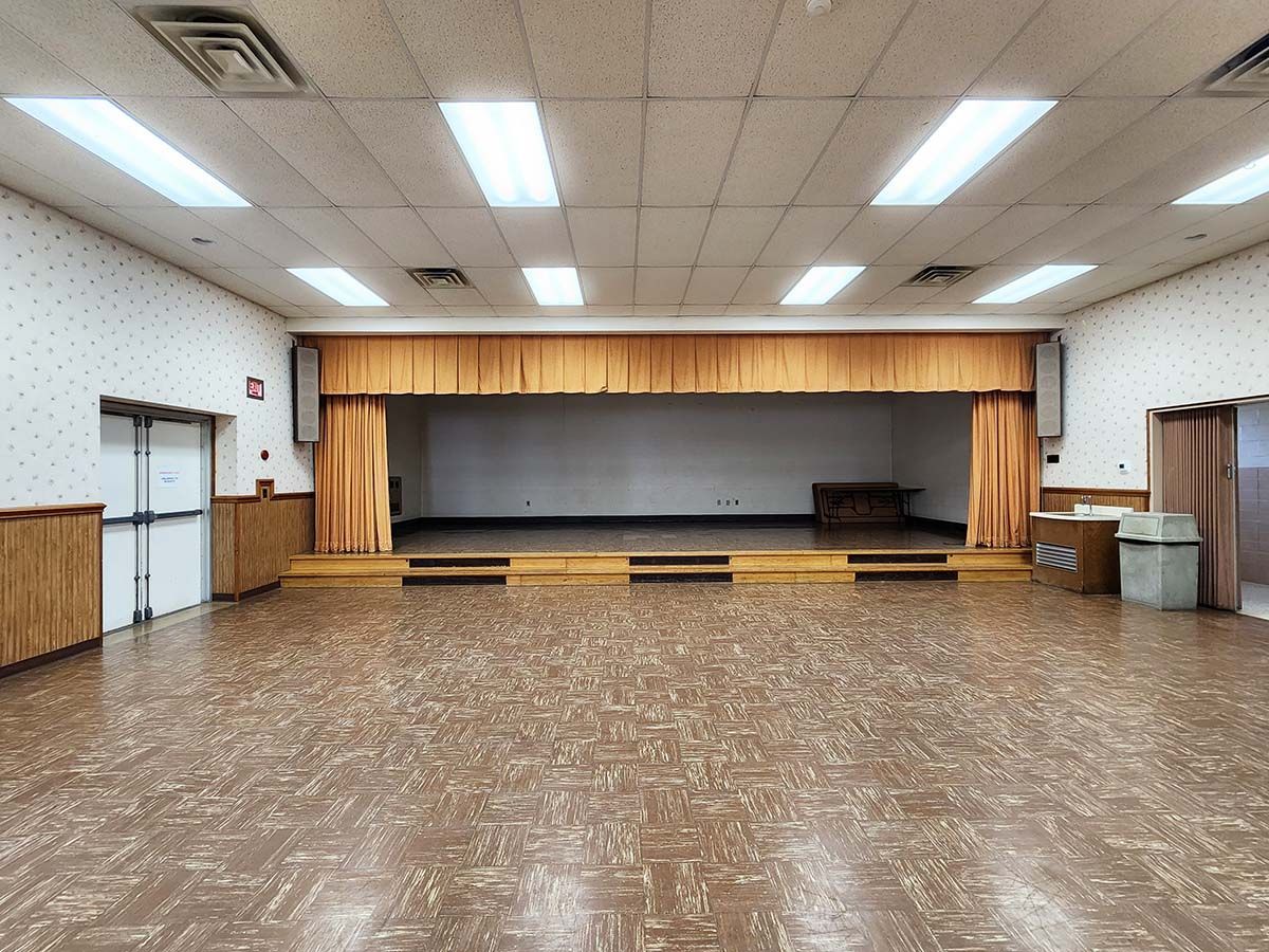 Empty community hall with a small stage, yellow curtains, and fluorescent ceiling lights
