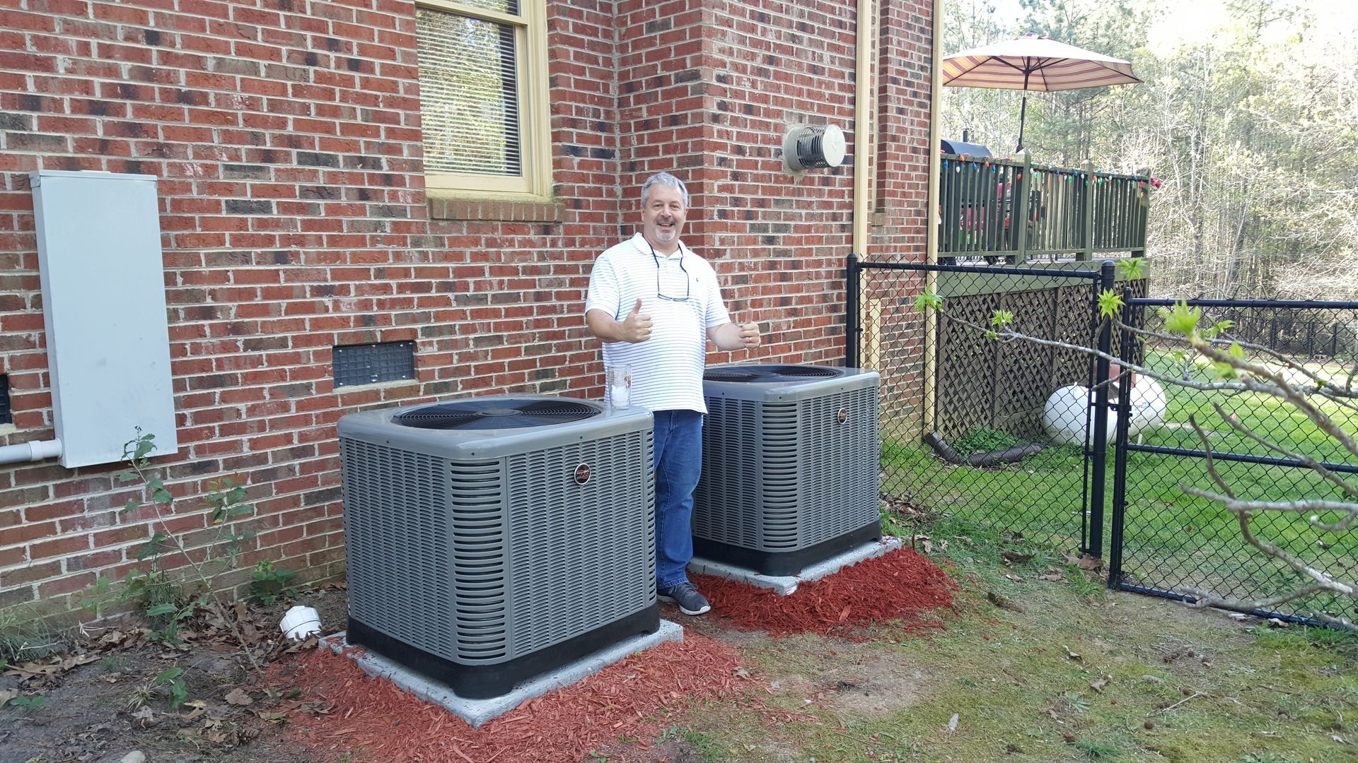 Man standing beside two outdoor HVAC units against a brick house wall