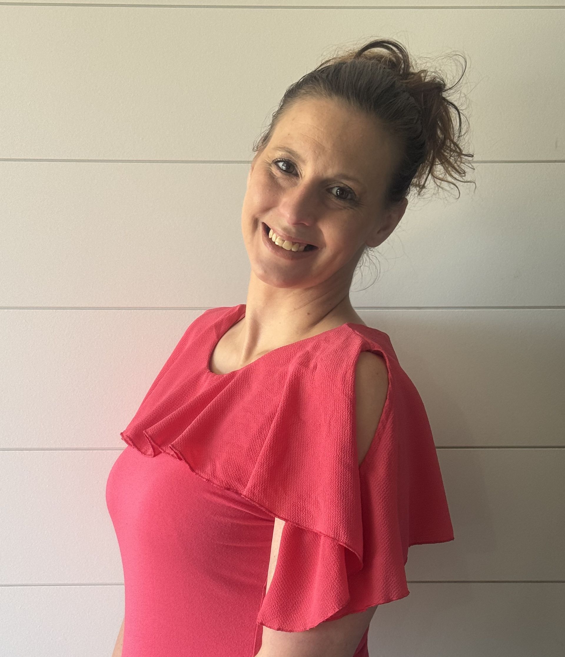 Woman in a pink top with shoulder cutouts, smiling in front of a white wall.