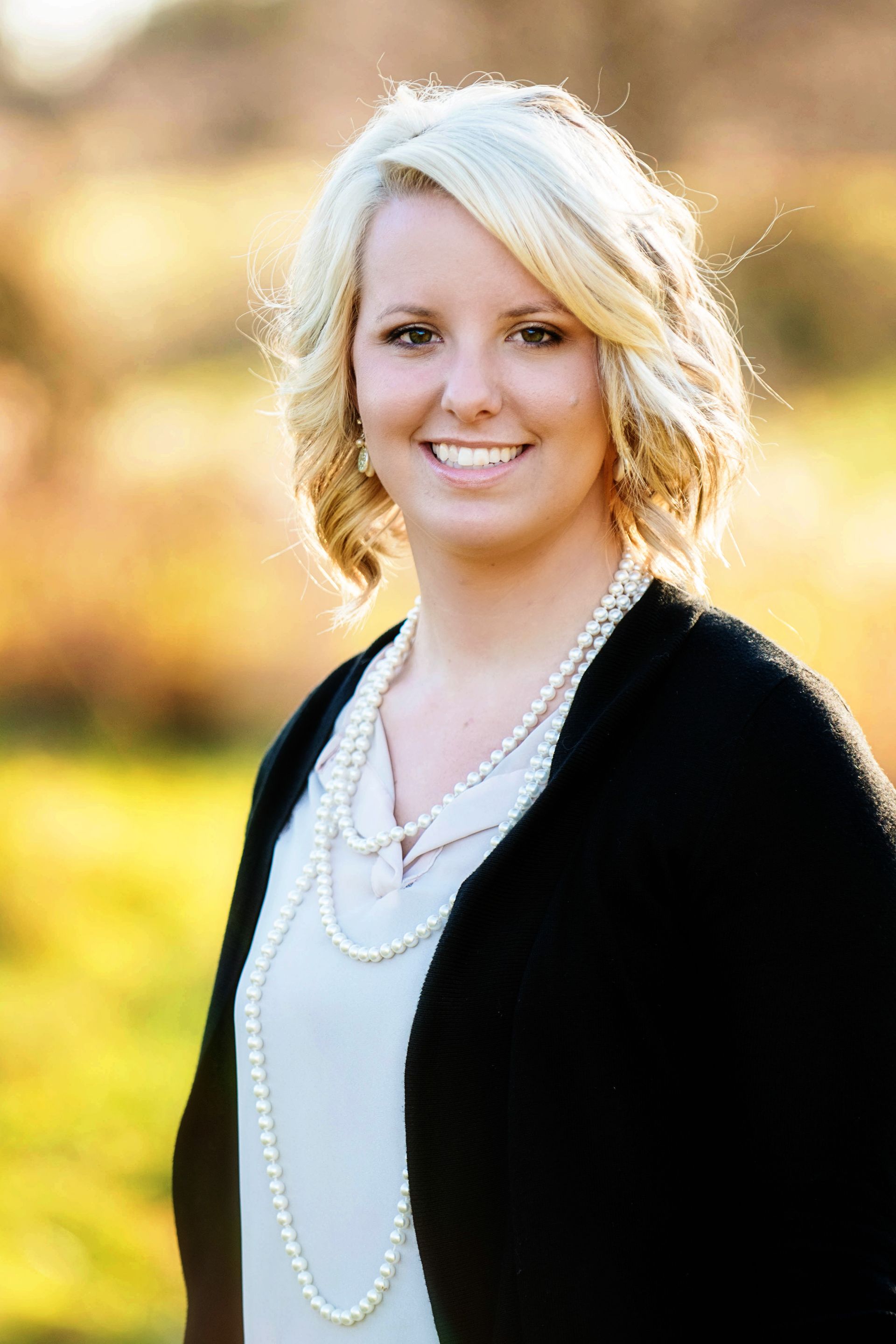Woman with blonde hair wearing a pearl necklace and black cardigan, smiling outside.