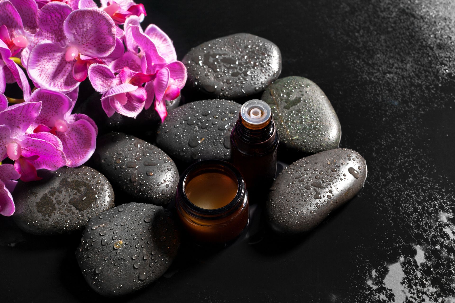 Candle burning in a brown glass jar on a wooden tray with essential oils, brush, and fabric; soft lighting.