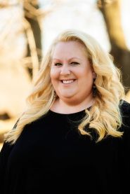 Woman with long blonde hair smiles, wearing a black top outdoors.