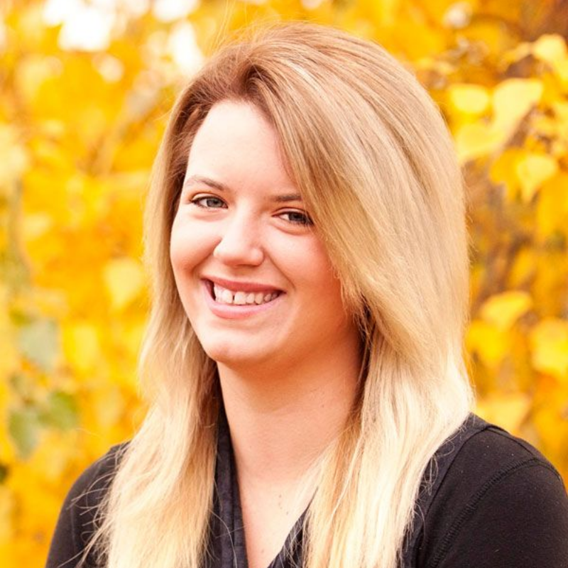 Woman with blonde hair smiling in front of yellow fall foliage. Wearing a black top.
