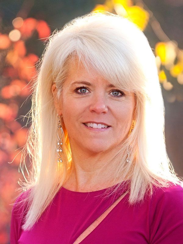Woman with blonde hair wearing a magenta top, smiling outdoors. Autumn foliage in background.