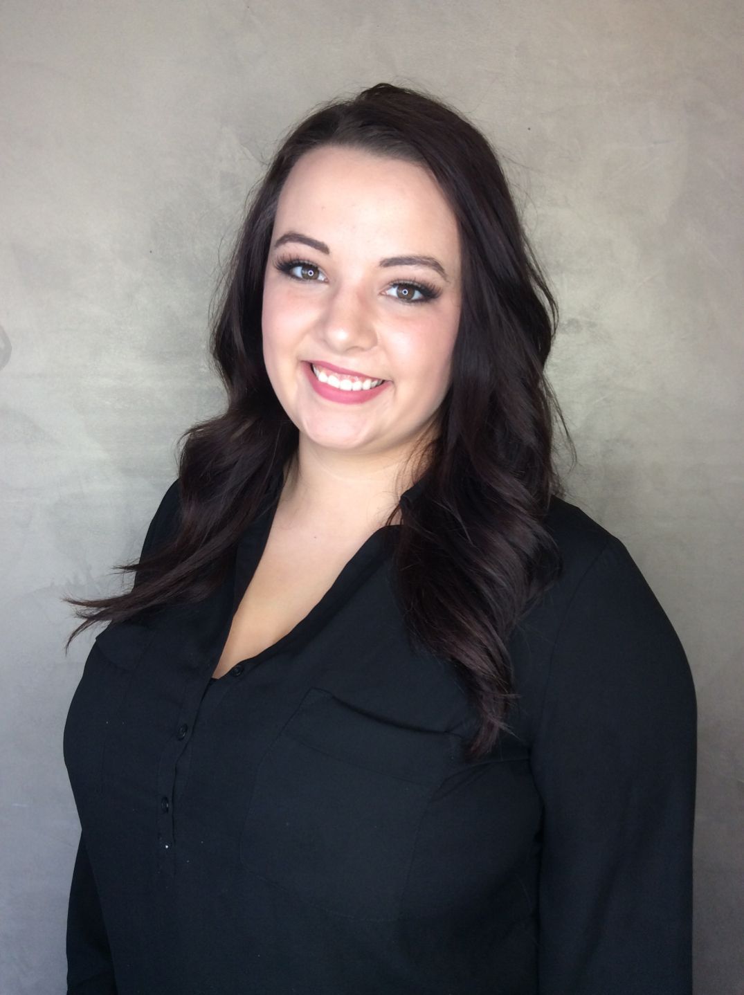 Woman with dark hair smiles, wearing a black blouse, posed against a gray background.