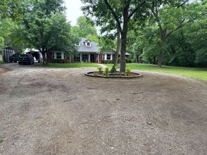 Gravel driveway with a tree island leading to a house surrounded by lush green trees